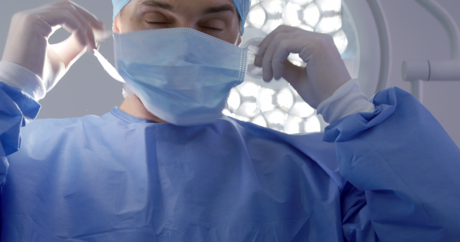 Caucasian male surgeon  healthcare professional in a hospital operating theatre wearing a surgical cap and gown, putting on a surgical mask. Healthcare workers in the Coronavirus Covid19 pandemic Royalty-Free Stock Footage #1043121772