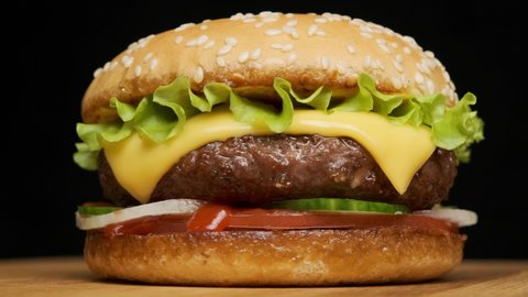 tasty beef burger with cheese and golden bun rotating on wooden table on black background.