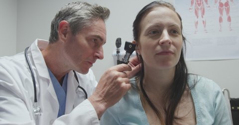 Side view of a Caucasian male healthcare professional wearing a lab coat and a stethoscope, examing the ear of a Caucasian female patient using an otoscope during a routine medical check up