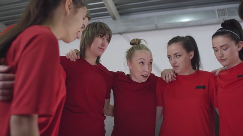 Team of young female soccer athletes in sports uniform standing in circle, embracing and listening to leader while getting ready for match in locker room