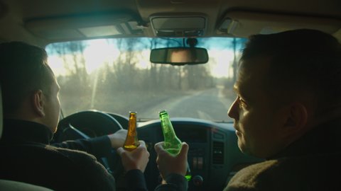 Scene of two men ride in a car with bottles of alcohol, they drink alcohol while driving, chatting, laughing. The camera is behind. Drunk driver idea and concept