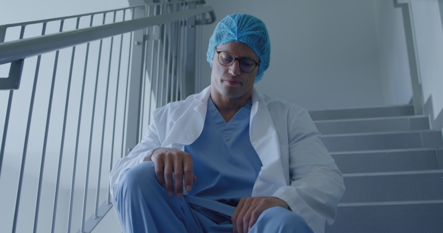 Front view of a troubled Caucasian male healthcare worker in a hospital, wearing glasses, a surgical cap and lab coat, sitting on the stairs. Healthcare workers in the Coronavirus Covid19 pandemic Royalty-Free Stock Footage #1043129749