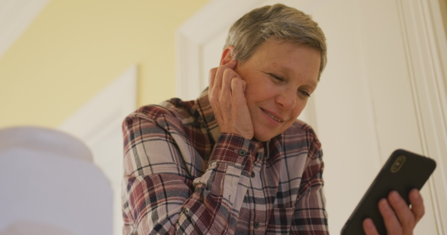 Low angle front view of a senior Caucasian woman at home, standing on the landing, leaning on the banister using a smartphone and smiling, slow motion