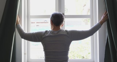 Rear view of a senior Caucasian woman with short hair standing and drawing the curtains at home, silhouetted against the window, slow motion,social distancing and self isolation in quarantine lockdown