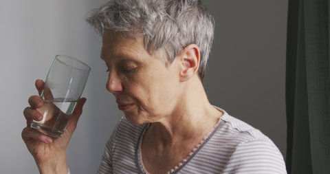 Side view close up of a senior Caucasian woman with short grey hair sitting, holding a glass of water and wiping her nose with a tissue in her bedroom at home, slow motion, social distancing 