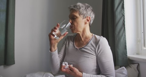 Side view of a senior Caucasian woman with short grey hair sitting on her bed and drinking a glass of water and holding a tissue in front of the window in her bedroom at home, slow motion