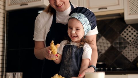 A little happy girl helps her mother knead the dough at home in the kitchen, she has all the palms in the dough. A cute child in a cap and apron is playing with dough in the kitchen.