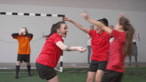 Group of young joyous female players jumping, huddling and giving high five to each other while celebrating soccer goal during match on indoor sports field