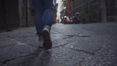 Low-angle handheld shot of a youthful woman walking down a dark old abandoned alleyway at dusk. Ground view of a woman's feet as she walks along a cobblestone path in Naples, Italy. Slow Motion, 4K.