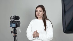 A woman blogger, in a white jumper, with long hair is recording video in a professional studio on a camera and an audio recorder. She speaks emotionally and makes hand gestures. Gray background