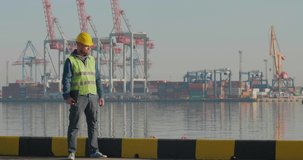 Docker With A Beard In A Yellow Helmet Stands With A Tablet PC In The Seaport  Against The Background Of Cranes And Containers. The Foreman Inspects The Industrial Harbor. Cinema 4K Video