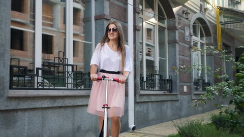 4k footage of beautiful smiling woman in pink skirt riding on city street using scooter