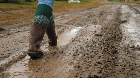 Feet in brown rubber boots goes through dirt country road and rain puddles. Man walking muddy road.