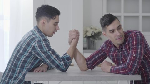 Close-up of two Caucasian men competing in arm-wrestling. Brunette man winning his twin brother. Siblings having fun at home. Leisure, resting, lifestyle.