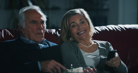 Close up of a happy senior couple is enjoying watching a movie on TV and eating a popcorn on sofa in living room at home.