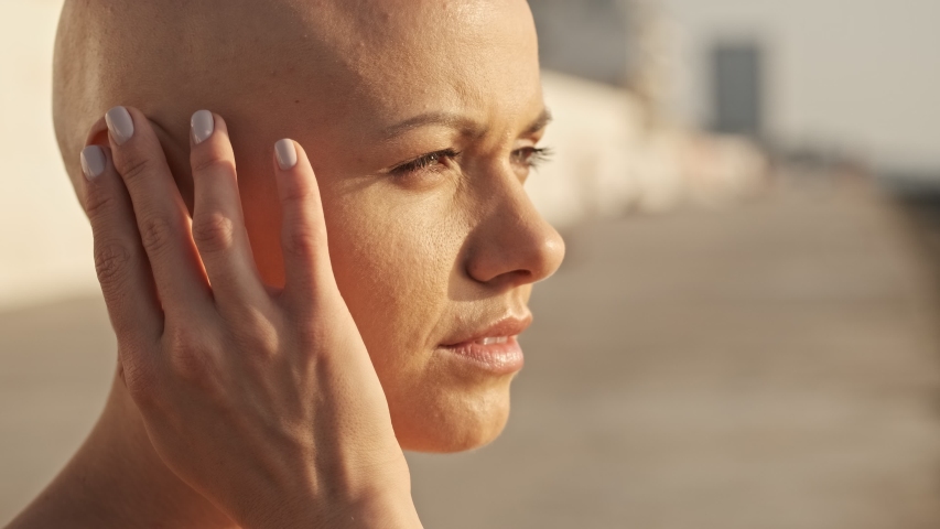 Side view of Pleased attractive bald sports woman in earphones listening music and looking away while sitting near the sea outdoors