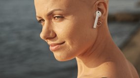 Side view of Pleased attractive bald sports woman in earphones listening music with closed eyes while sitting near the sea outdoors