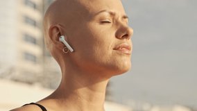 Side view of Pleased attractive bald sports woman touching earphone and becoming happy while sitting near the sea outdoors