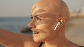 Close up view of Smiling attractive bald sports woman in earphones with finger shadow on the face listening music and looking away while sitting near the sea outdoors