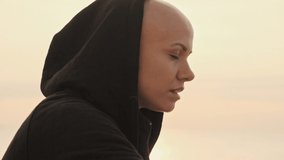 Side view of smiling attractive bald sports woman in hood looking away and enjoying this moment near the sea outdoors