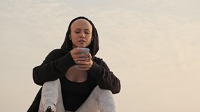 Smiling attractive bald sports woman in hood and earphones using smartphone while sitting near the sea outdoors