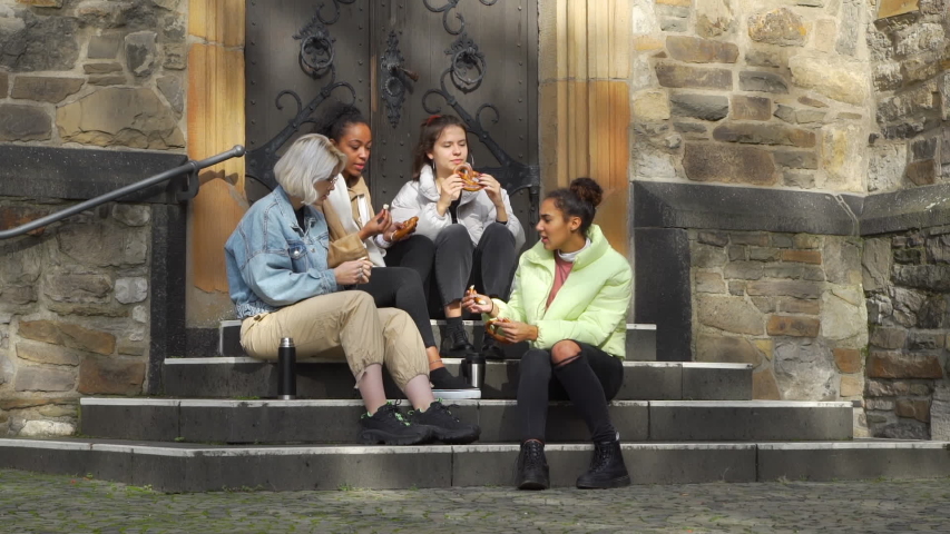 Four Multiracial Female Teens Eating Pretzels and Smiling while Sitting on the Steps of Ancient European Building. Street Food and Fast food Concept