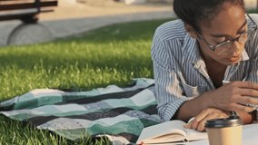Calm pretty african woman in eyeglasses studying with book while lying on plaid in the park