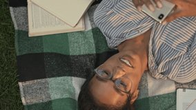 Top view of Cheerful pretty african woman in eyeglasses using smartphone while lying on plaid in the park