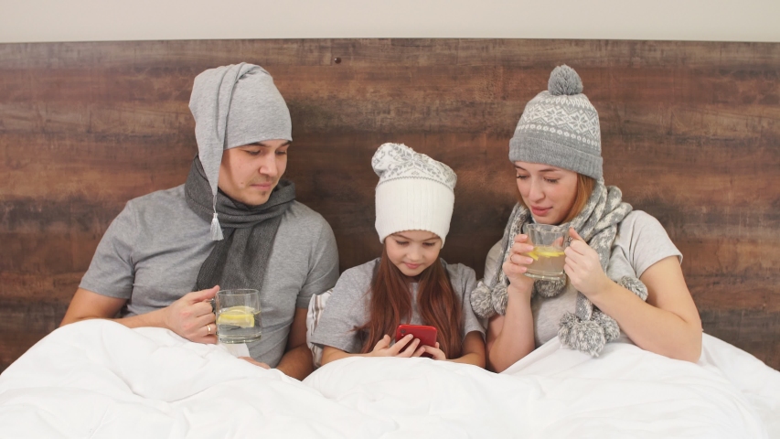 Sick family lying on bed together, cute kid girl lying between mother and father and hold smartphone in hands
