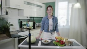 home blog, modern attractive housewife writes vlog video on mobile phone, talks about healthy foods and losing weight, shows diet plan while standing at table with fresh vegetables in kitchen