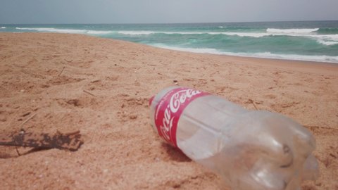 Umdloti, KwaZulu-Natal, South Africa 12/2/2019 Static hand held shot of a discarded coca cola bottle and other natural beach rubbish, waves crashing, blue hazy sky, golden sand, illustrative editorial