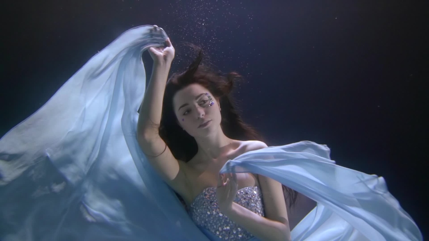 a beautiful girl with long flowing hair and a sequined dress is floating under the water. she waves her arms and blue cloth like wings
