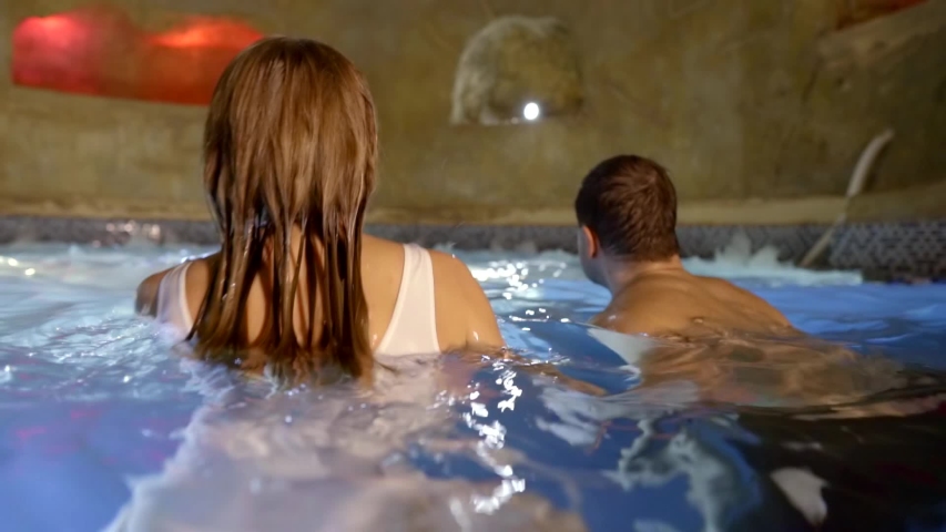 a man and a woman swim in a small round grotto and come out of the water. the view from the back