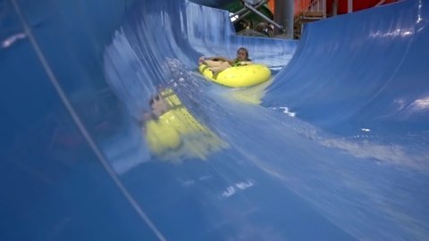 A little girl is in a water Park, she slides down a blue slide on a yellow tubing, drives into the water and spray flies. She's wearing a green inflatable vest.