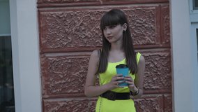 Happy female teen in light green dress drinks beverage in blue paper cup on street and easily poses with smile on face at the building with windows. 4K