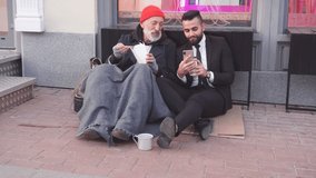 Rich man in black suit sit with phone, show interesting videos while beggar eating and talking.