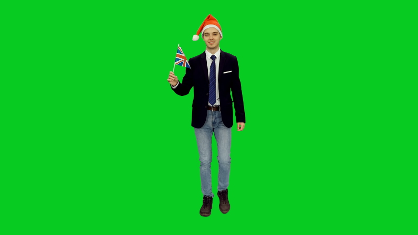 Friendly young man in santa hat walking with waving flag of UK against green screen background, Front view, Full body, 4k chroma key footage