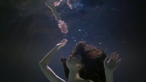 a girl with long dark flowing hair floats face up underwater. she touches her reflection on the surface of the water with her palms and removes her hands