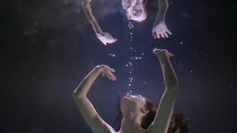 Close-up portrait of a face of a dark-haired girl with rhinestones on her face, who is underwater on a blue background, she is reflected from the surface of the water, pulling her hands up.