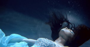 A close-up portrait of a pretty dark-haired girl with rhinestones on her face, who is underwater on a blue background, the delicate fabric of her dress is developing, bubbles are floating around her.
