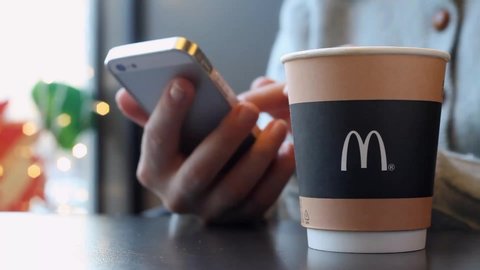MINSK, BELARUS - December 19, 2019: Paper cup of coffee with McDonald's logo in hands of a woman closeup. Woman drinks a coffee and uses a smartphone on McDonald's Restaurant