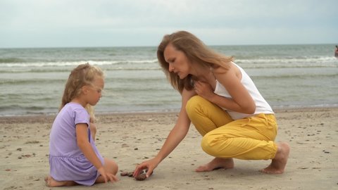 Mother with little daughter playing with pebble stones on the beach. Caucasian family girls on background of sea. Camera motion shot with gimbal