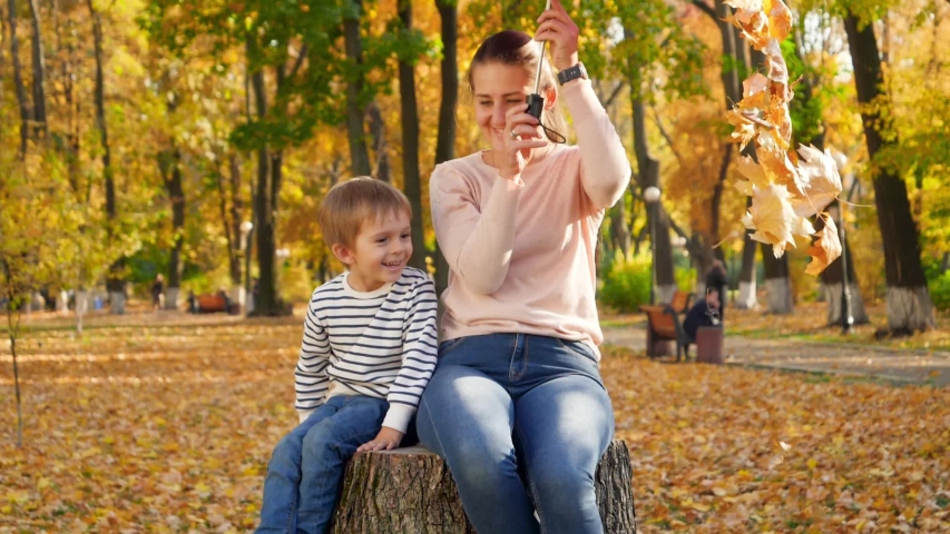 Slow motion footage of golden autumn leaves falling from umbrella on young mother and smiling little boy sittin on tree stump at park