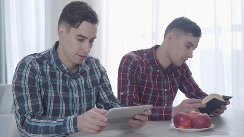Portrait of adult Caucasian man playing video game on tablet and showing it to his twin brother. Brunette guy reading book and looking at device screen. Siblings resting indoors.