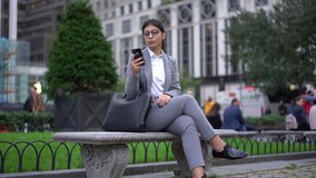 Cheerful spanish millennial female manager in formal wear and glasses checking banking account via smartphone and 4G internet connection sending notification on street