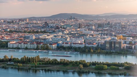 Aerial panoramic view of Vienna city with skyscrapers, historic buildings and a riverside promenade timelapse in Austria. Evening skyline before sunset from Danube Tower viewpoint. Mountains on a