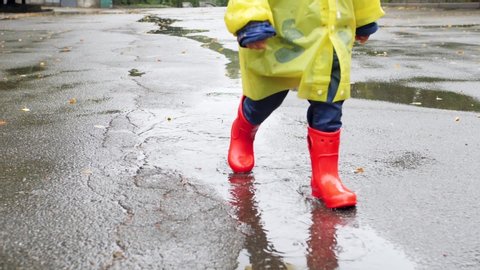 Slow motion footage of little boy in red wellington boots and yellow raincoat walking over big puddles and splashing water after big rain in park