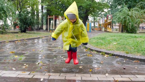 4k slow motion footage of cheerful little boy in raincoat and wellington boots jumping in puddle and splashing water at autumn park during rain