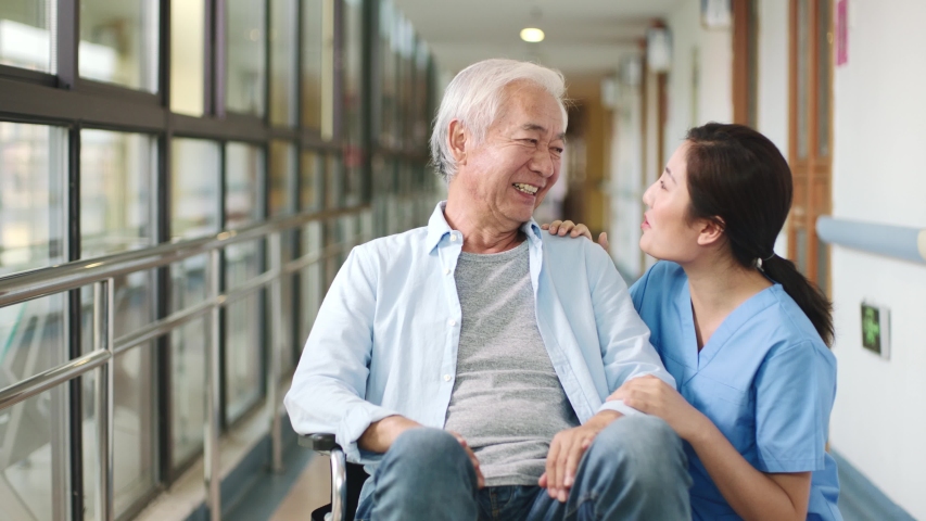 friendly asian worker talking to wheelchair bound senior man resident in hallway of assisted living facility Royalty-Free Stock Footage #1043314204
