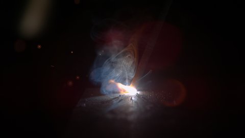 SLOW MOTION, MACRO, DOF: Round weld glows in the dark workshop as an unrecognizable metalworker welds a piece of metal. Sparks fly off a craftsman's welding torch as he welds an iron workpiece.